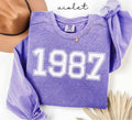 Purple (violet) sweatshirt with '1987' printed on it, worn by a person, with a plain background.