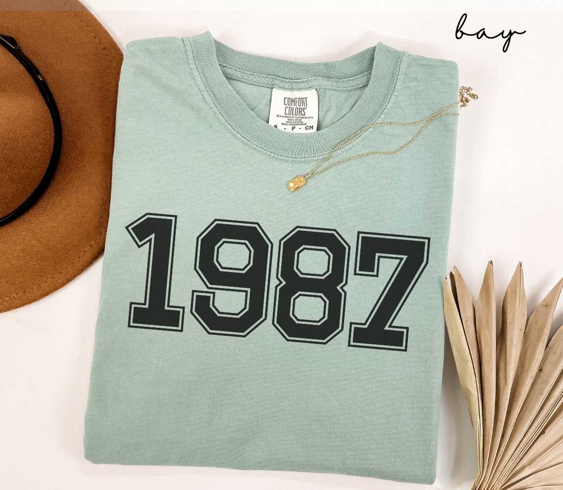 A light green (bay) t-shirt with "1987" printed on it, placed on a white surface alongside a hat and dried leaves.

