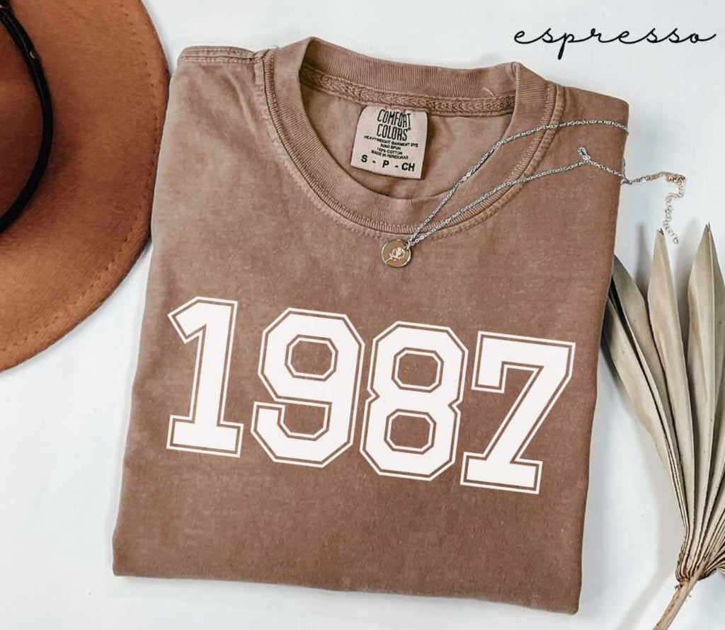 Folded brown (espresso) t-shirt with '1987' print on a white surface, accompanied by a hat and dried leaves.
