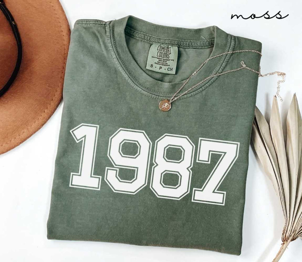 Folded green (moss) t-shirt featuring a '1987' print on a white surface alongside a hat and dried leaves.
