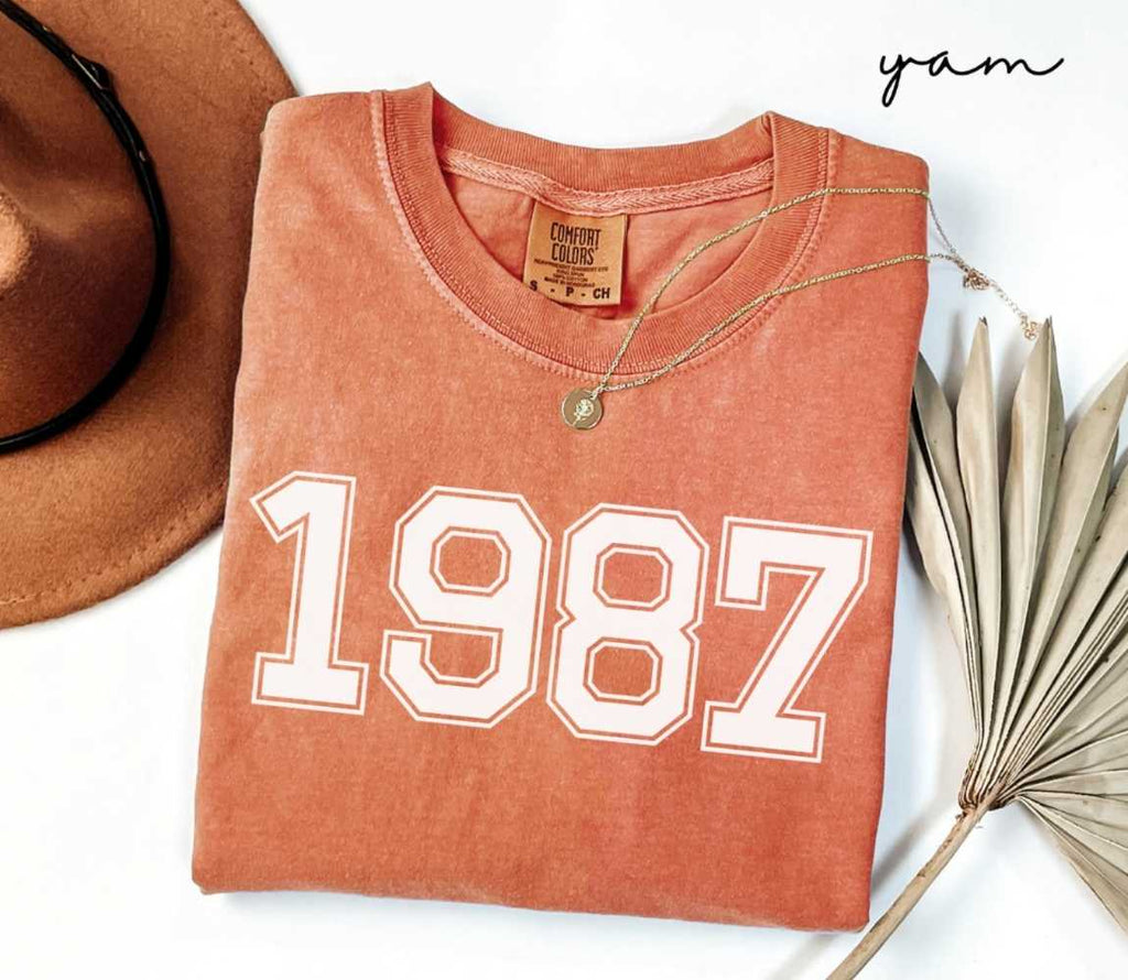 Folded orange (yam) t-shirt with a '1987' print, a brown hat, and a dried plant on a white background.

