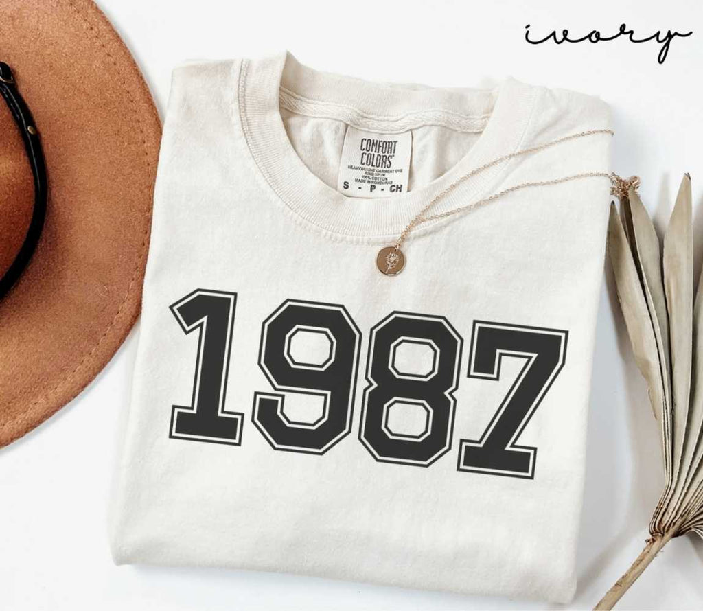 A beige (ivory) t-shirt with '1987' print, placed next to a brown hat and a dried plant on a light background.
