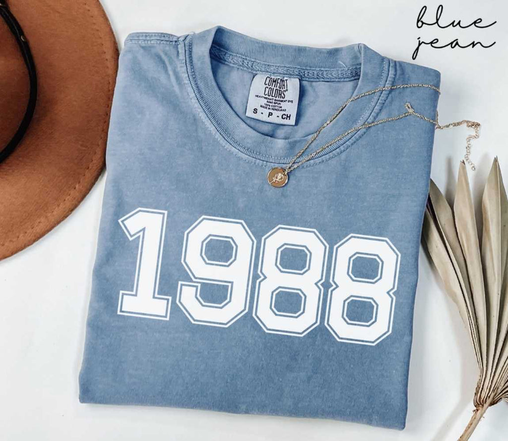 Folded blue jean t-shirt featuring '1988' print on a white surface, accompanied by a hat and dried leaves.
