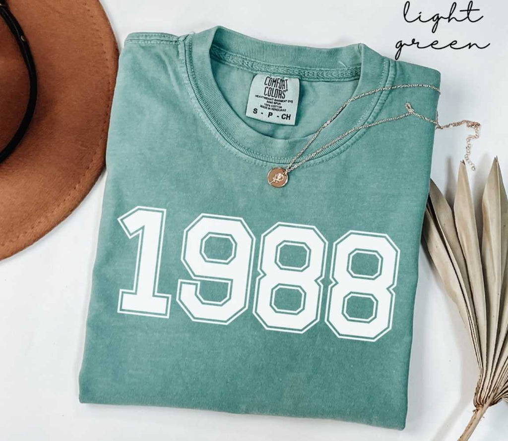 A light green t-shirt with '1988' printed on it, placed on a white surface, accompanied by a hat and dried leaves.

