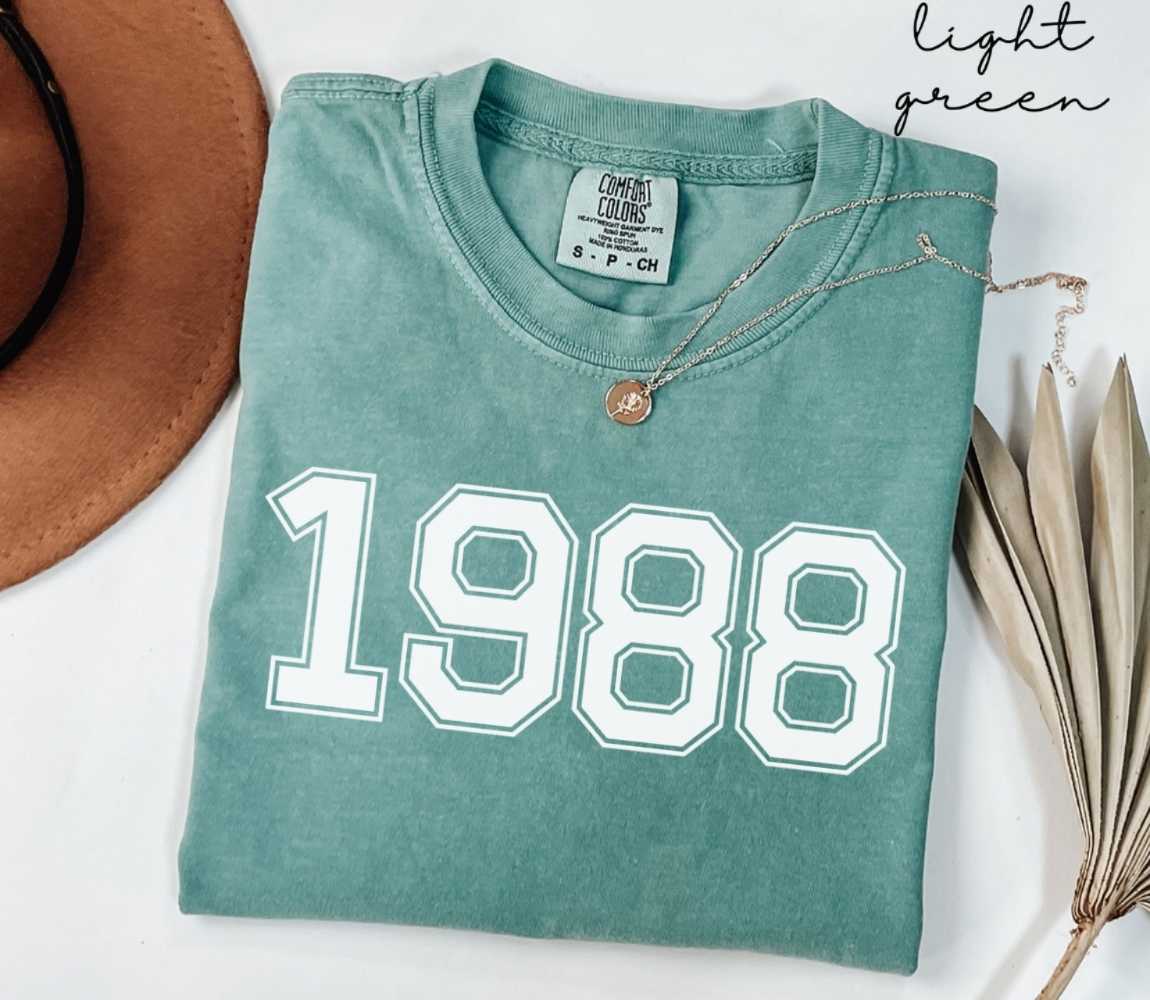 A light green t-shirt with '1988' printed on it, placed on a white surface, accompanied by a hat and dried leaves.
