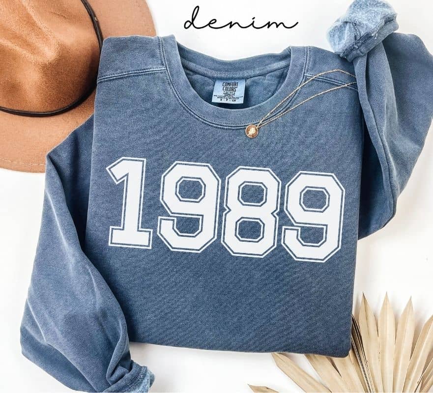 Blue (denim) sweatshirt with '1989' printed on it, worn by a person, with a plain background.