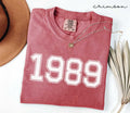 A red (Crimson) folded t-shirt with '1989' printed on it, next to a hat, and dried leaves on a white background.
