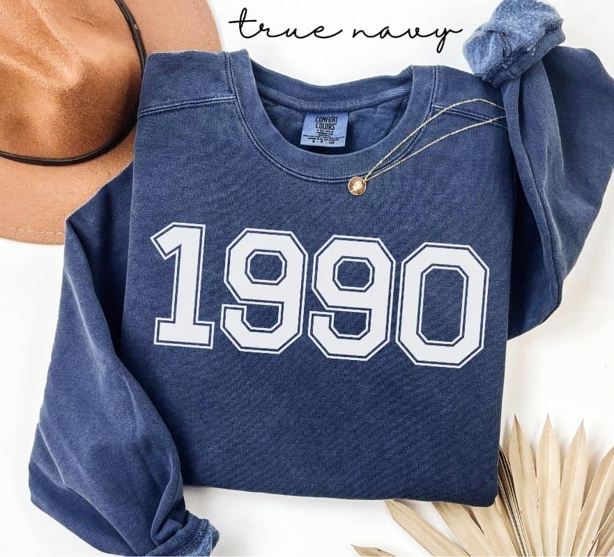 Blue (true navy)  sweatshirt with '1990' printed on it, displayed with a hat and decorative elements.