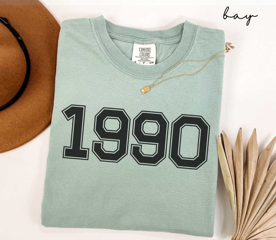 A light green (bay) t-shirt with "1990" printed on it, placed on a white surface alongside a hat and dried leaves.
