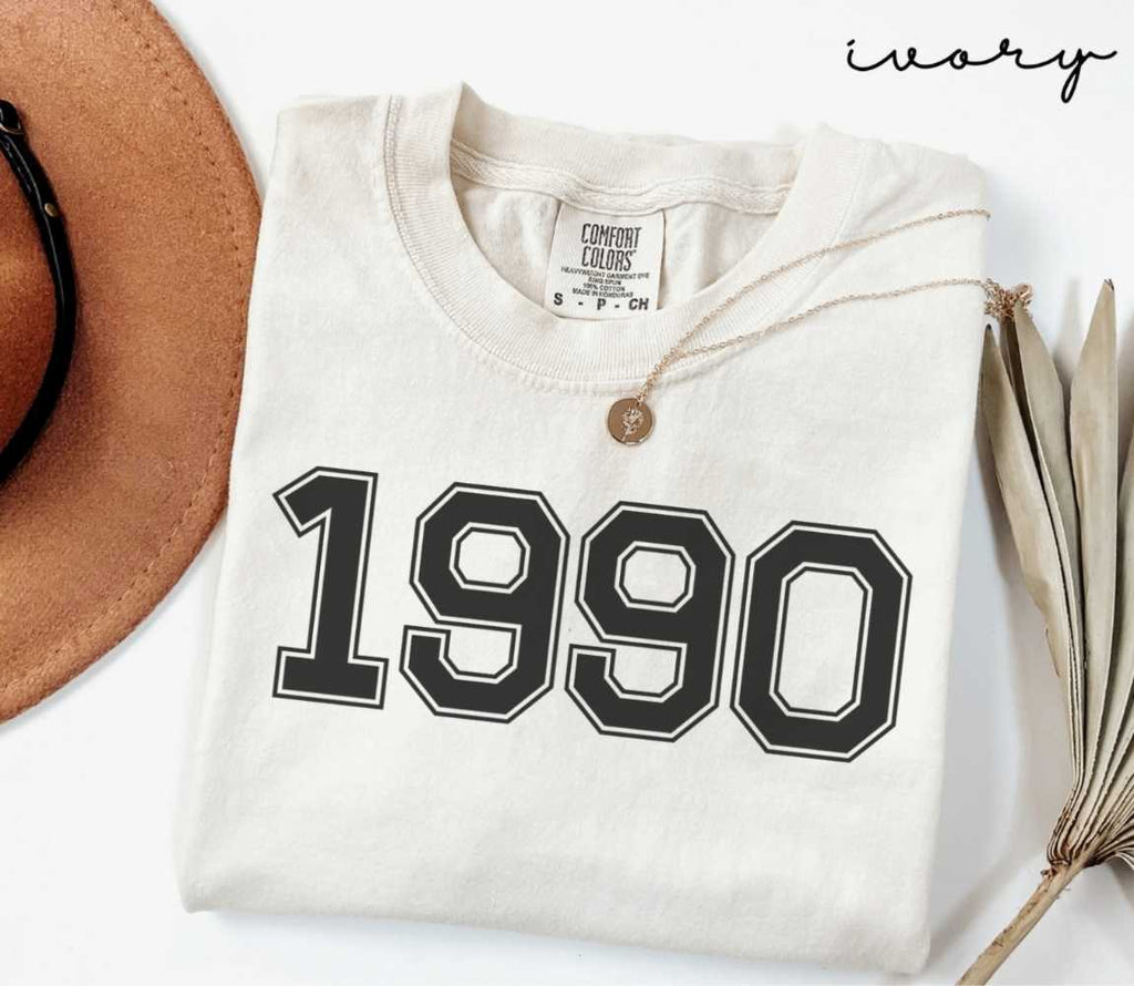 A beige (ivory) t-shirt with '1990' print, placed next to a brown hat and a dried plant on a light background.
