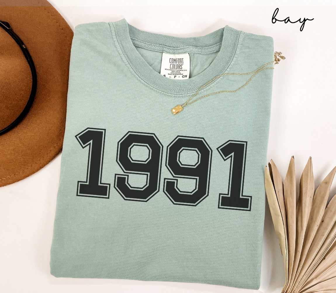 A light green (bay) t-shirt with "1991" printed on it, placed on a white surface alongside a hat and dried leaves.
