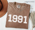 Folded brown (espresso) t-shirt with '1991' print on a white surface, accompanied by a hat and dried leaves.
