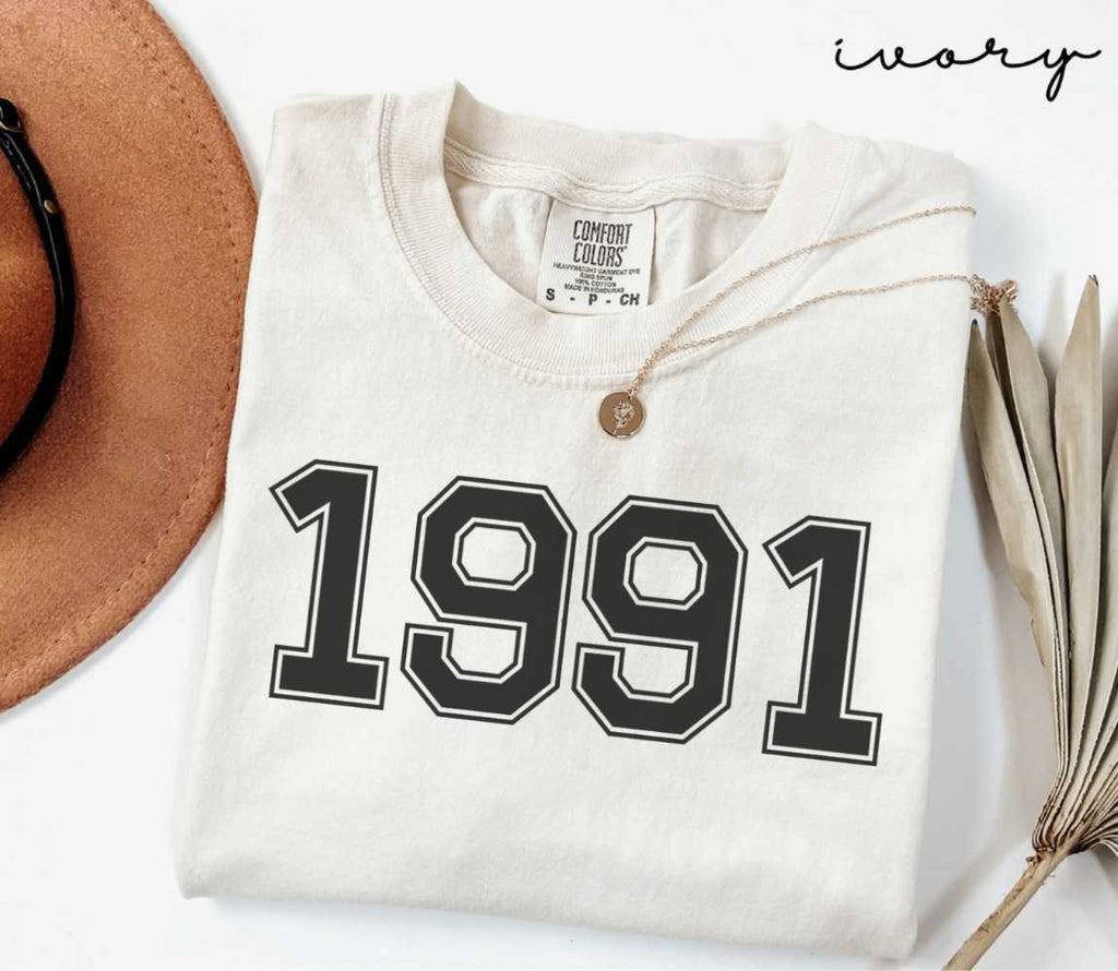A beige (ivory) t-shirt with '1991' print, placed next to a brown hat and a dried plant on a light background.
