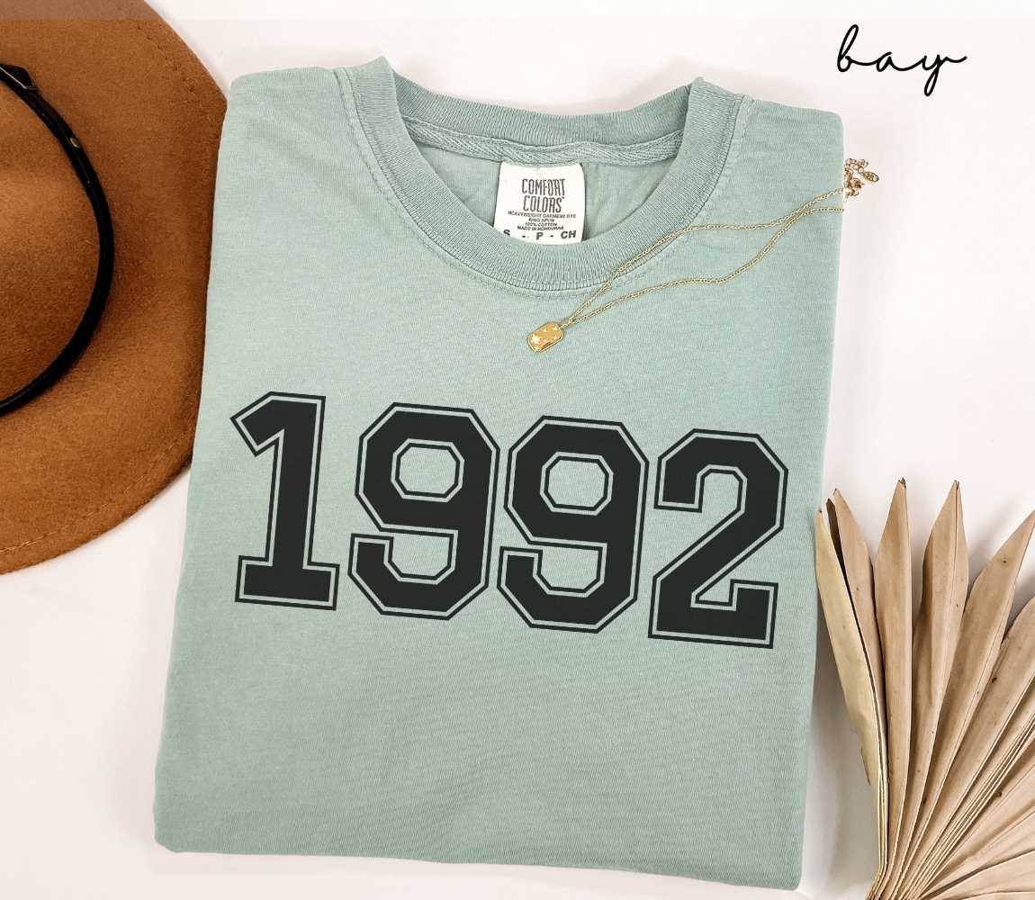 A light green (bay) t-shirt with "1992" printed on it, placed on a white surface alongside a hat and dried leaves.

