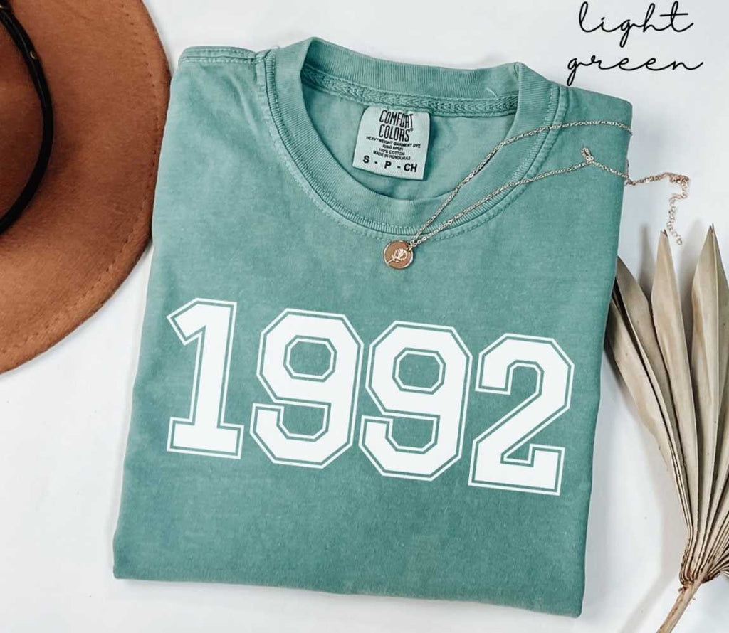 A light green t-shirt with '1992' printed on it, placed on a white surface, accompanied by a hat and dried leaves.
