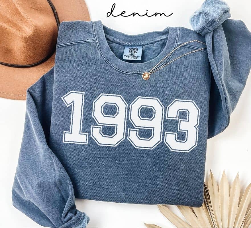 Blue (denim) sweatshirt with '1993' printed on it, worn by a person, with a plain background.
