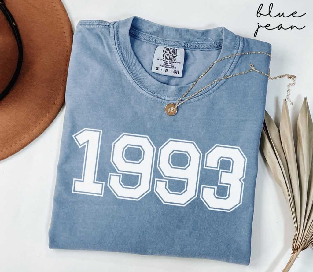 Folded blue jean t-shirt featuring '1993' print on a white surface, accompanied by a hat and dried leaves.
