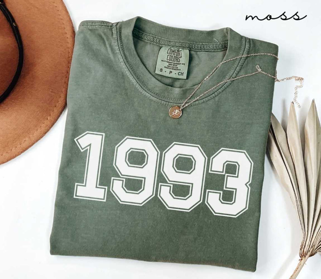 Folded green (moss) t-shirt featuring a '1993' print on a white surface alongside a hat and dried leaves.
