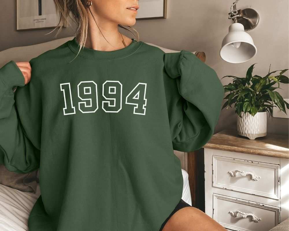 A person is sitting in a bedroom, wearing a forest green sweatshirt that has '1994' printed on it.
