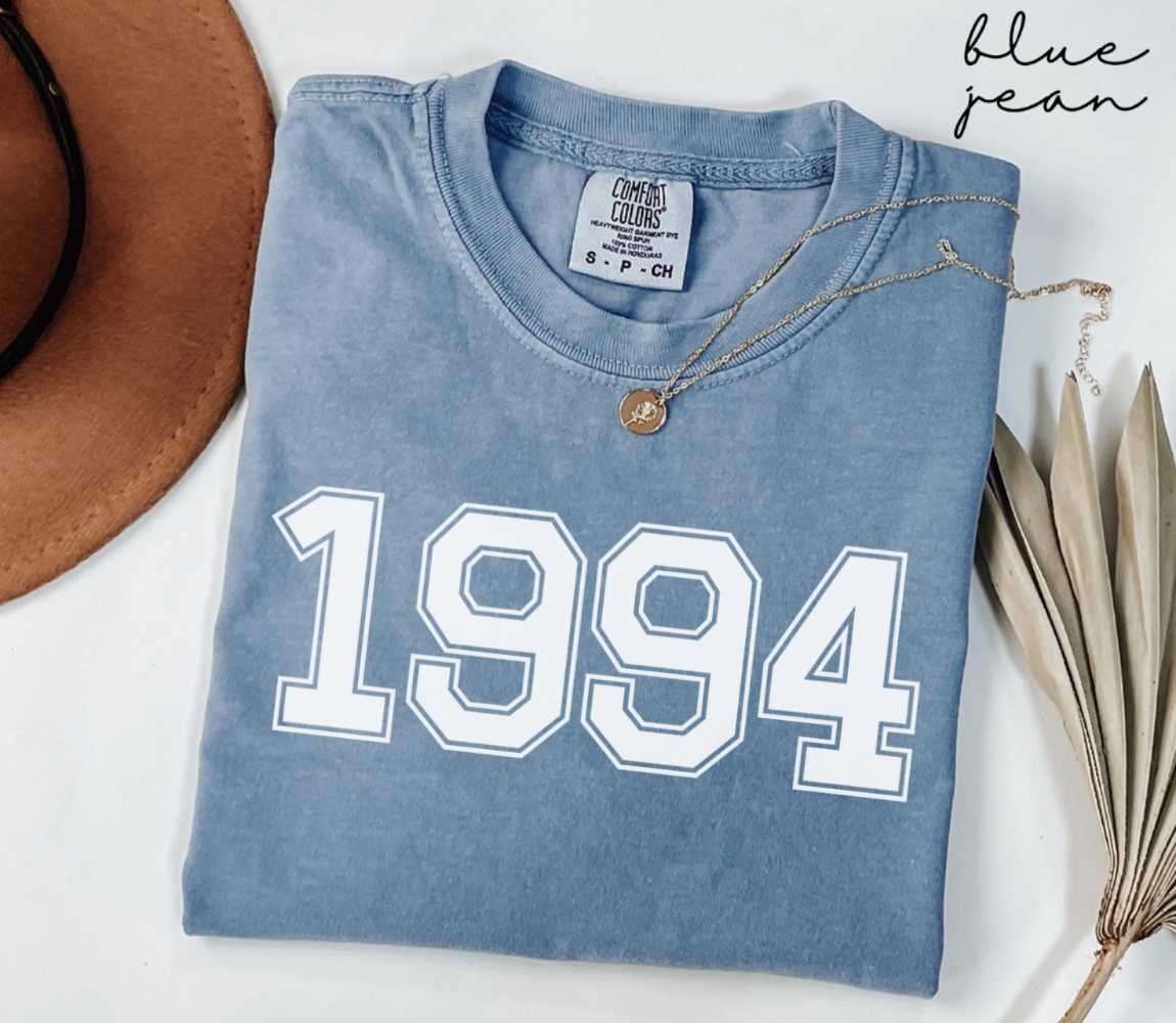 Folded blue jean t-shirt featuring '1994' print on a white surface, accompanied by a hat and dried leaves.
