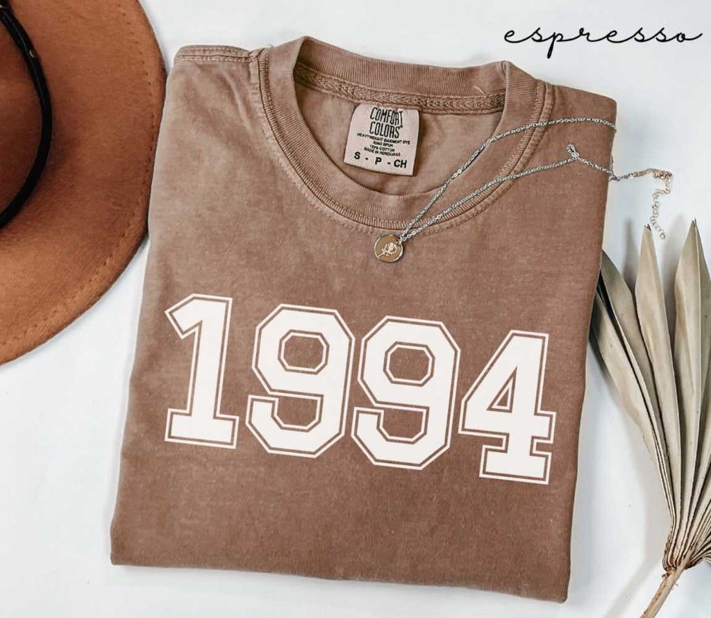 Folded brown (espresso) t-shirt with '1994' print on a white surface, accompanied by a hat and dried leaves.
