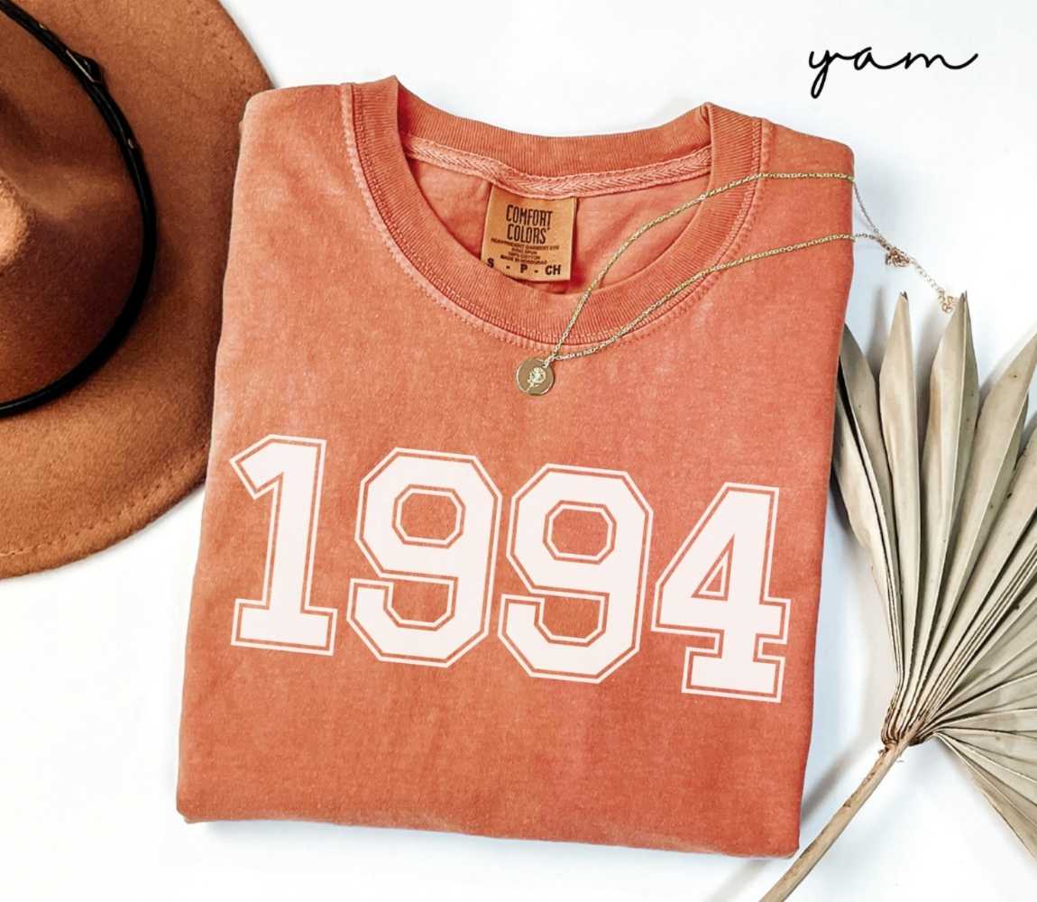 Folded orange (yam) t-shirt with a '1994' print, a brown hat, and a dried plant on a white background.
