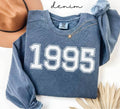 Blue (denim) sweatshirt with '1995' printed on it, worn by a person, with a plain background.
