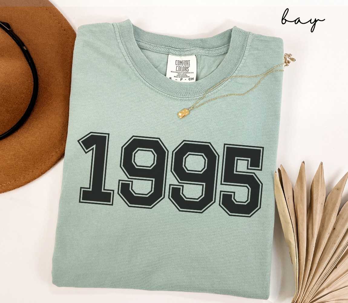 A light green (bay) t-shirt with "1995" printed on it, placed on a white surface alongside a hat and dried leaves.
