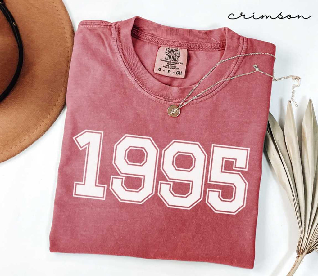 A red (Crimson) folded t-shirt with '1995' printed on it, next to a hat, and dried leaves on a white background.
