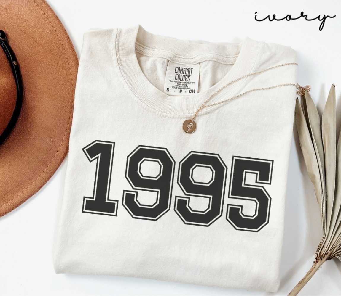 A beige (ivory) t-shirt with '1995' print, placed next to a brown hat and a dried plant on a light background.
