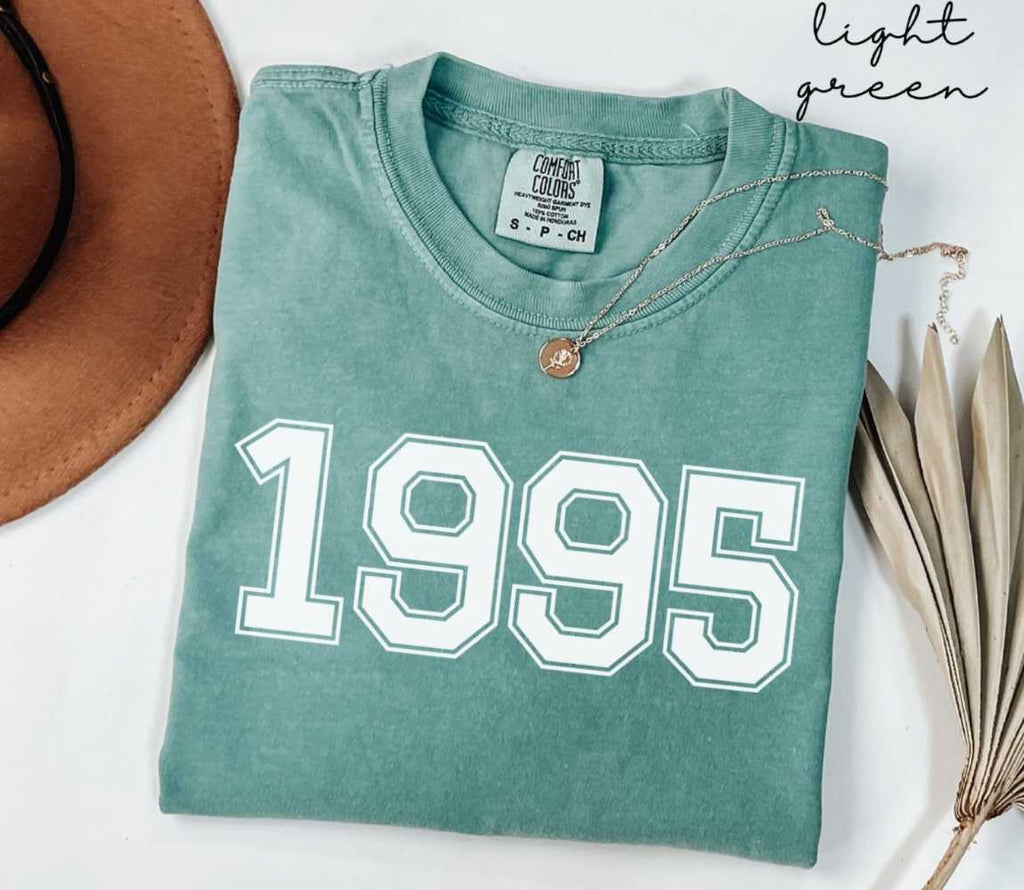 A light green t-shirt with '1995' printed on it, placed on a white surface, accompanied by a hat and dried leaves.
