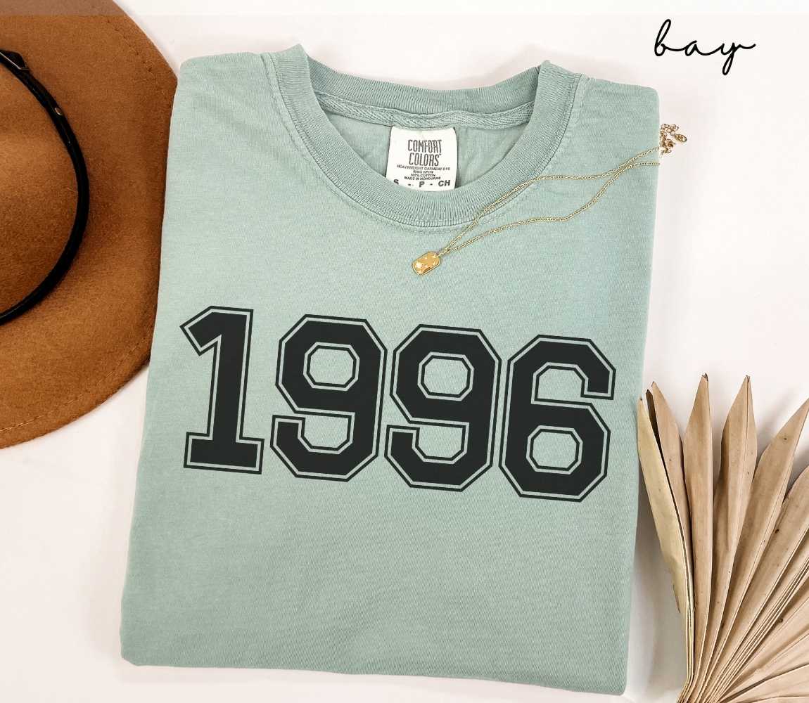 A light green (bay) t-shirt with "1996" printed on it, placed on a white surface alongside a hat and dried leaves.
