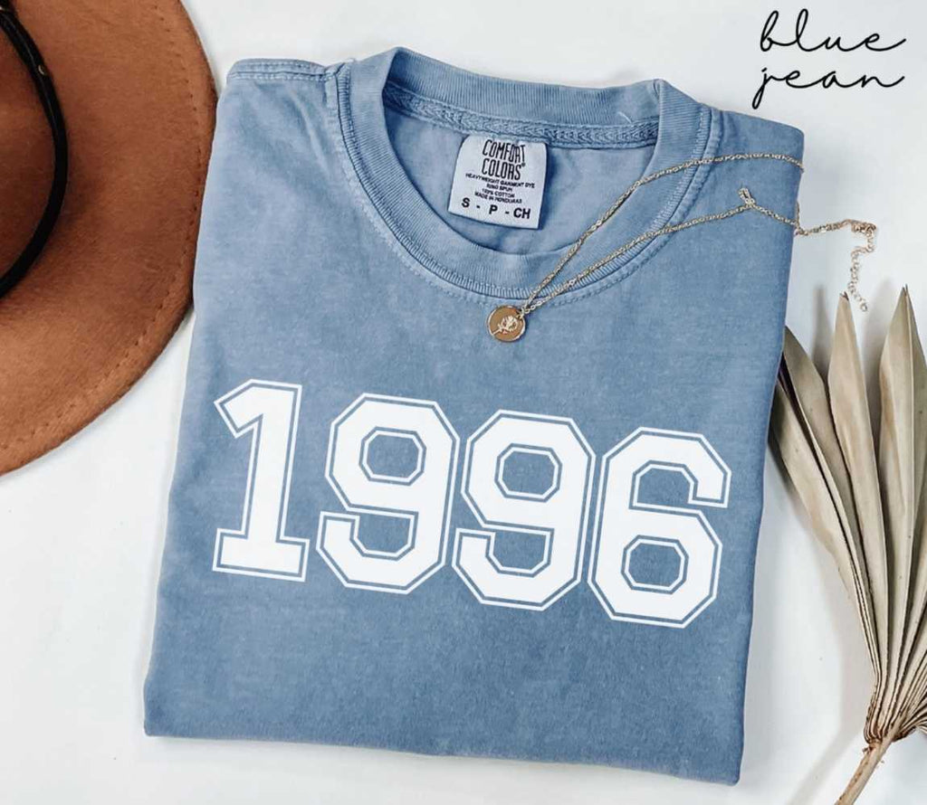 Folded blue jean t-shirt featuring '1996' print on a white surface, accompanied by a hat and dried leaves.
