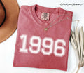 A red (Crimson) folded t-shirt with '1996' printed on it, next to a hat, and dried leaves on a white background.
