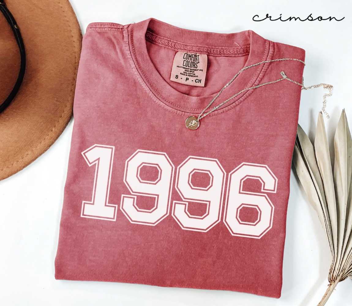 A red (Crimson) folded t-shirt with '1996' printed on it, next to a hat, and dried leaves on a white background.
