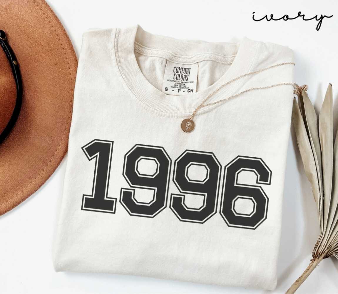 A beige (ivory) t-shirt with '1996' print, placed next to a brown hat and a dried plant on a light background.

