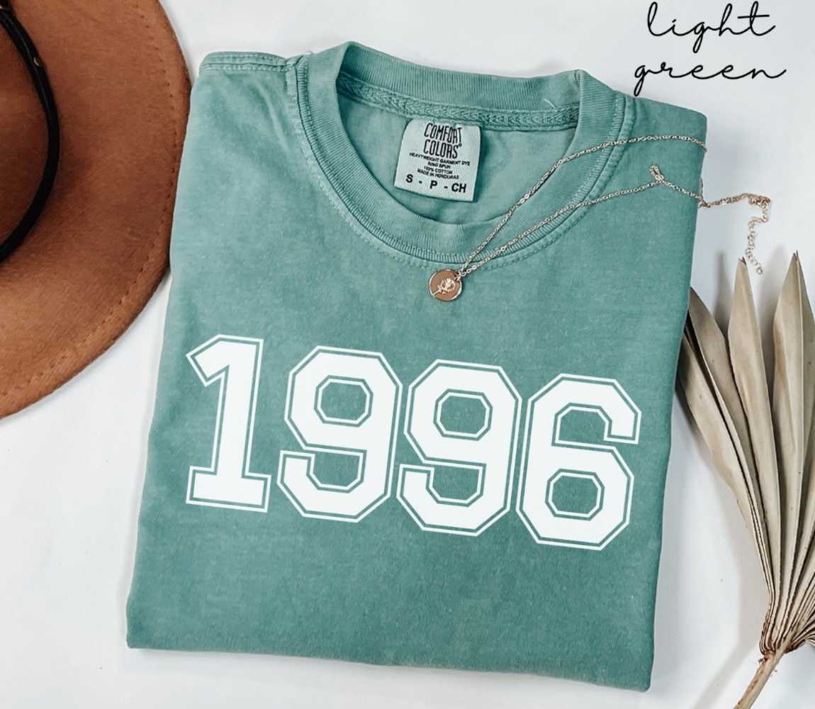 A light green t-shirt with '1996' printed on it, placed on a white surface, accompanied by a hat and dried leaves.
