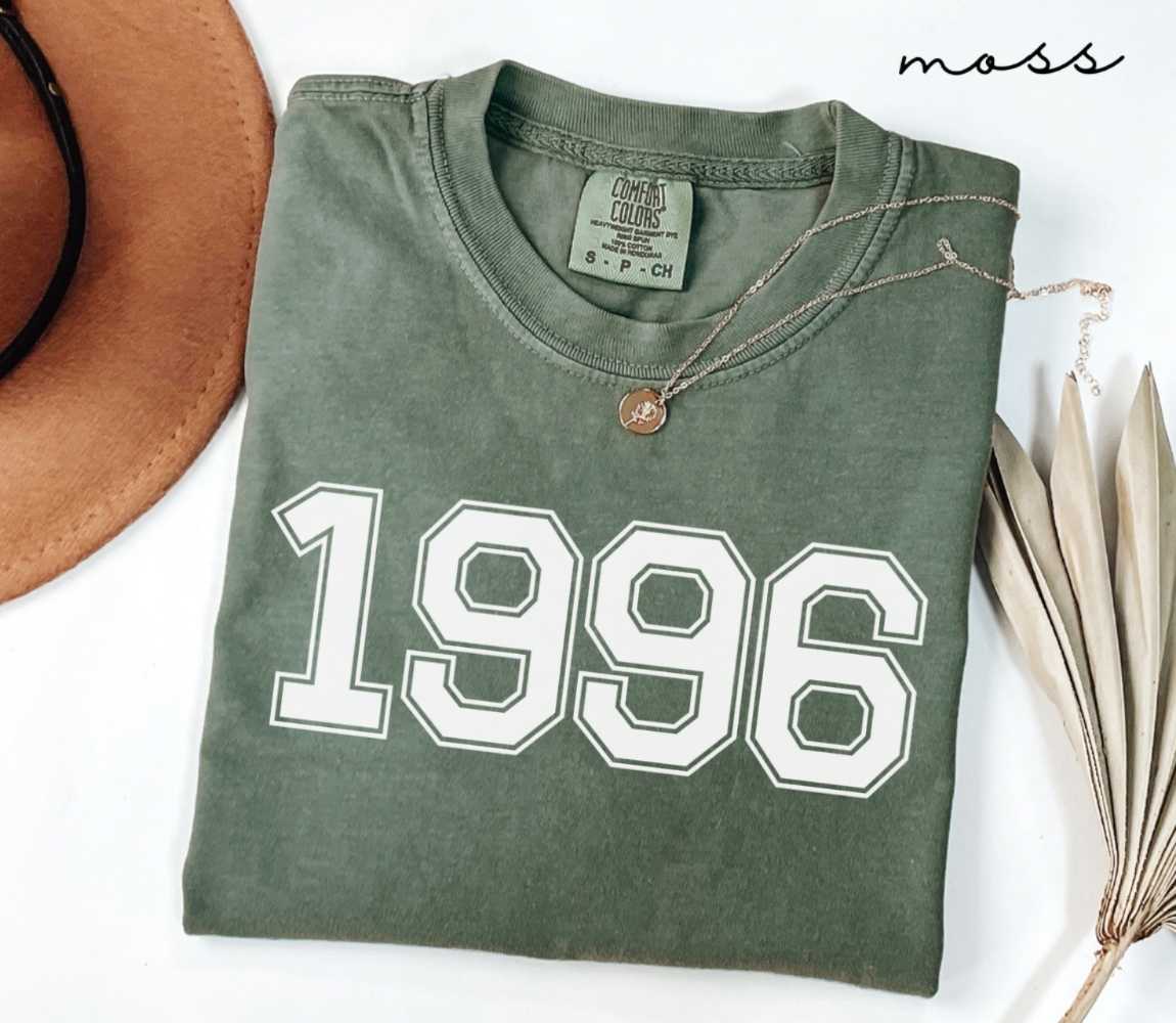Folded green (moss) t-shirt featuring a '1996' print on a white surface alongside a hat and dried leaves.
