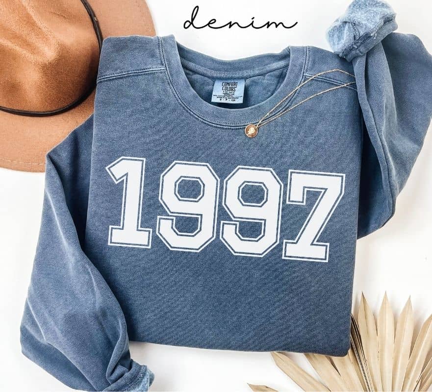 Blue (denim) sweatshirt with '1997' printed on it, worn by a person, with a plain background.
