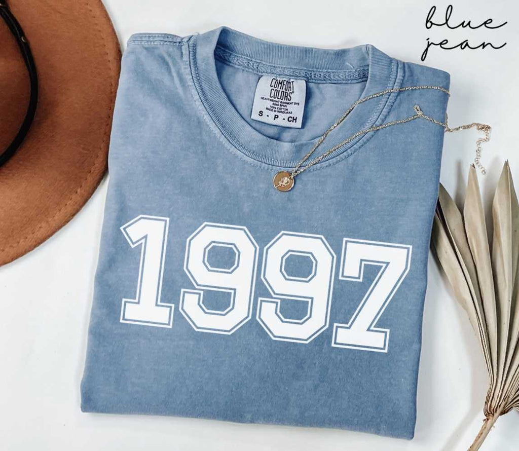 Folded blue jean t-shirt featuring '1997' print on a white surface, accompanied by a hat and dried leaves.
