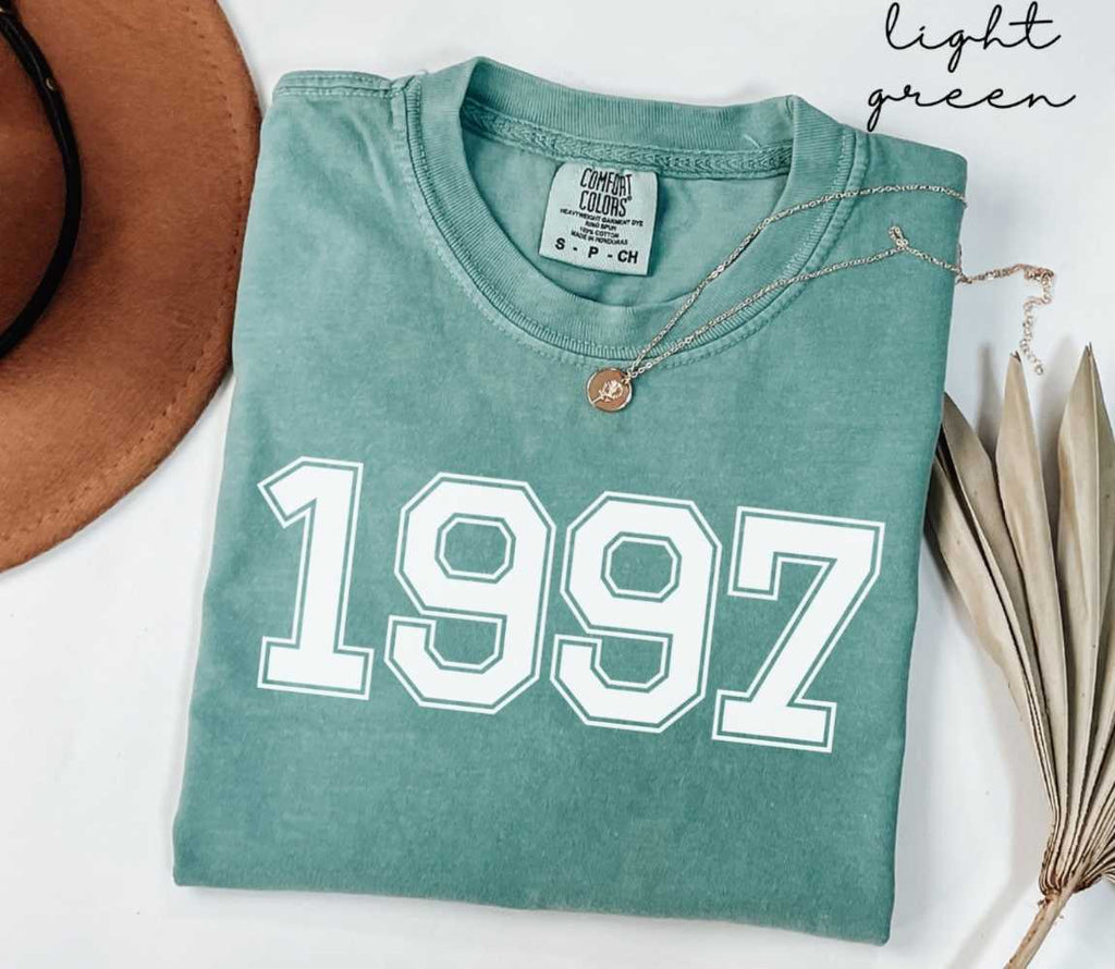 A light green t-shirt with '1997' printed on it, placed on a white surface, accompanied by a hat and dried leaves.
