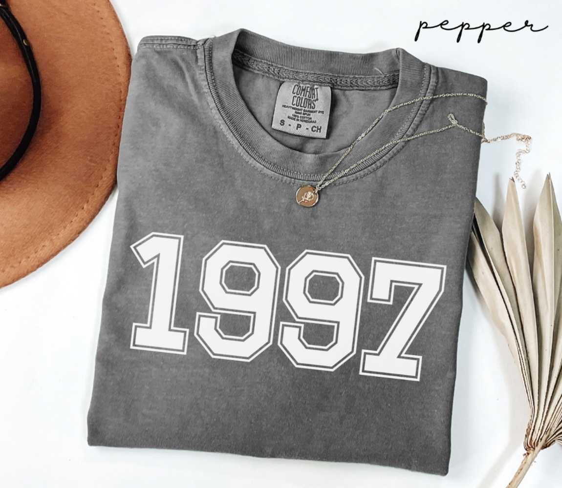 Folded gray (pepper) t-shirt with '1997' print on a white surface with a hat and dried leaves.
