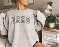 A person is sitting in a room wearing a gray (ash) sweatshirt with "1998" printed on it.
