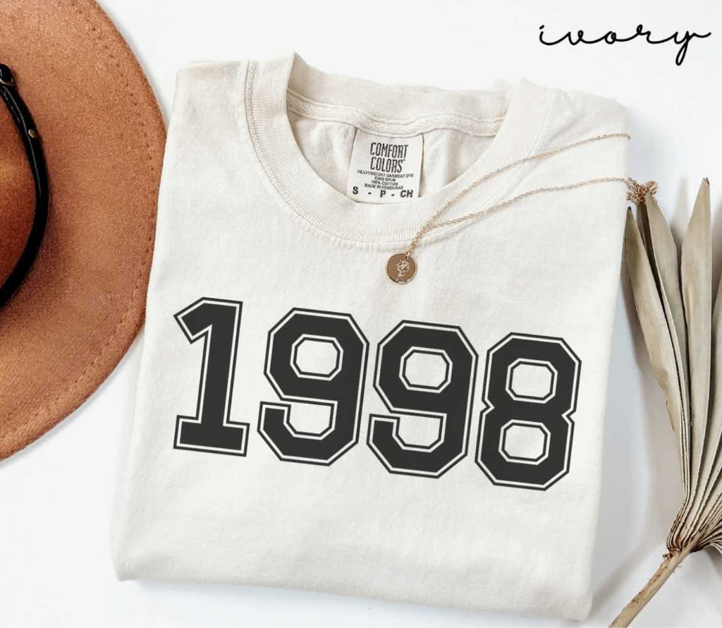 A beige (ivory) t-shirt with '1998' print, placed next to a brown hat and a dried plant on a light background.
