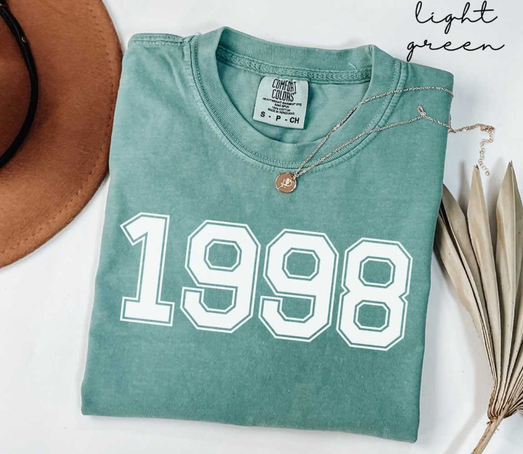 A light green t-shirt with '1998' printed on it, placed on a white surface, accompanied by a hat and dried leaves.
