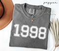 Folded gray (pepper) t-shirt with '1998' print on a white surface with a hat and dried leaves.
