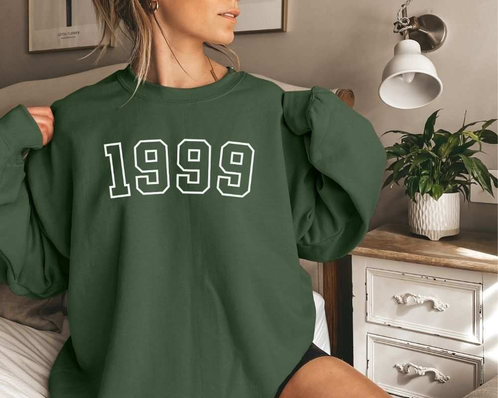 A person is wearing a forest green sweatshirt that has '1999' printed on it while standing in a bedroom.
