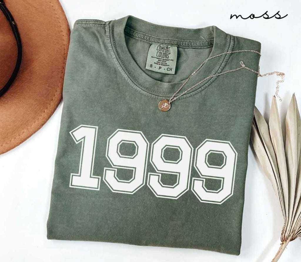 Folded green (moss) t-shirt featuring a '1999' print on a white surface alongside a hat and dried leaves.
