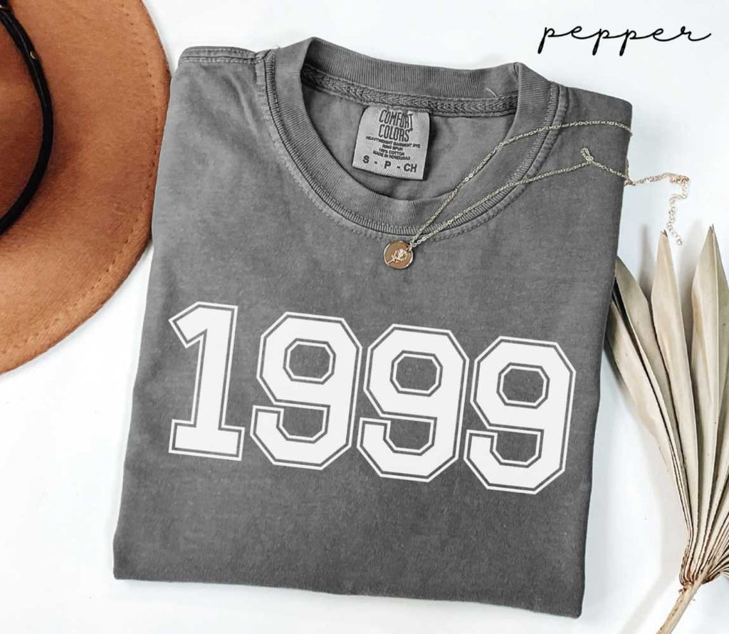 Folded gray (pepper) t-shirt with '1999' print on a white surface with a hat and dried leaves.
