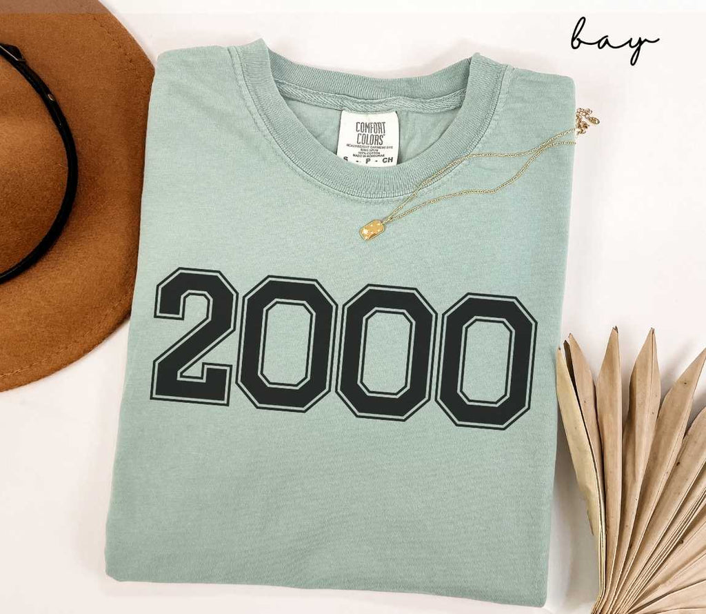 A light green (bay) t-shirt with "2000" printed on it, placed on a white surface alongside a hat and dried leaves.
